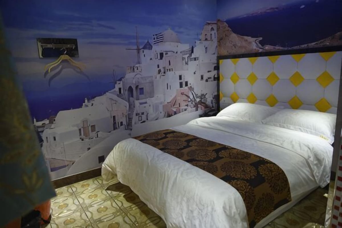A bedroom with a bed, a patterned rug, and a wall mural of a coastal town.