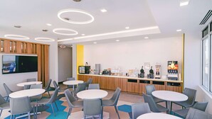 A modern cafe with round tables and chairs, a kitchen area with coffee machines, and a TV mounted on the wall. - Microtel Inn & Suites by Wyndham Newark NY (Newark)