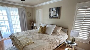 Ein Schlafzimmer mit einem großen Bett, einem Deckenventilator, einem Fenster mit Jalousien und einem kleinen Tisch mit Lampe.