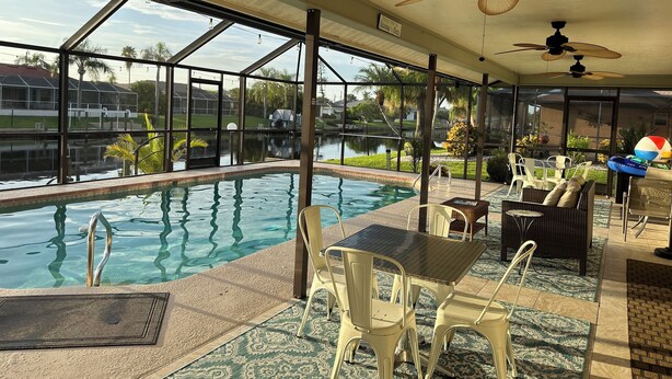Ein Innenpool mit freiem Blick auf die Außenwelt, darunter ein See und Wohnhäuser. Deckenventilatoren, Sitzgelegenheiten im Freien und ein Tisch am Pool sind vorhanden.