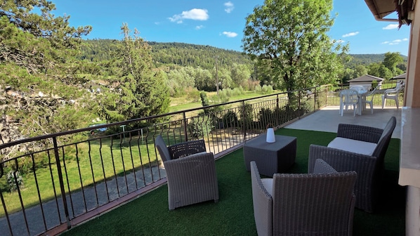 A balcony with wicker furniture and artificial grass, overlooking a forested landscape.