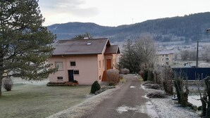 A two-story house with a sloped roof, a front porch, and a driveway leading to a road. - Apartment in quiet house between lakes and fir trees (MARTIGNAT)