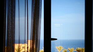 A view through a window with sheer curtains, revealing a coastal landscape with palm trees and a building.