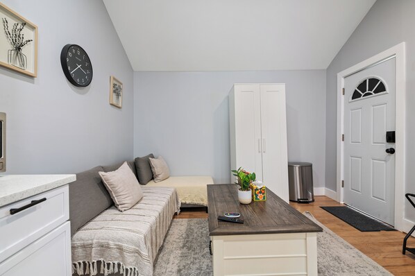 Una habitación pequeña y moderna con una cama, un sofá, una mesa de madera, un armario blanco y una puerta con ventana.