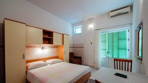 A bedroom with a bed, a table, a chair, a wardrobe, a window with blinds, and an air conditioning unit. - Apartments Pezo - Studio  ( Tina 3) (Baška Voda)