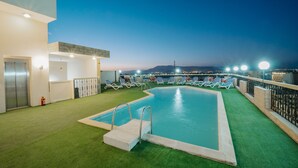 An indoor swimming pool with a view of the city at dusk, surrounded by lounge chairs and a building with a modern design. - Queens Valley Hotel (Luxor Governorate)