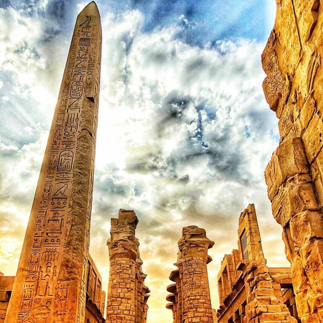Een eeuwenoude Egyptische obelisk met hiërogliefen, omringd door stenen ruïnes en kolommen.
