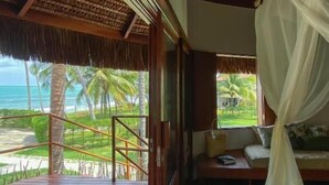 A room with a wooden deck, a bed with a canopy, and a view of the beach and palm trees.