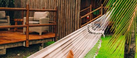 A hammock on a grassy lawn with a wooden deck and thatched-roof structures in the background.