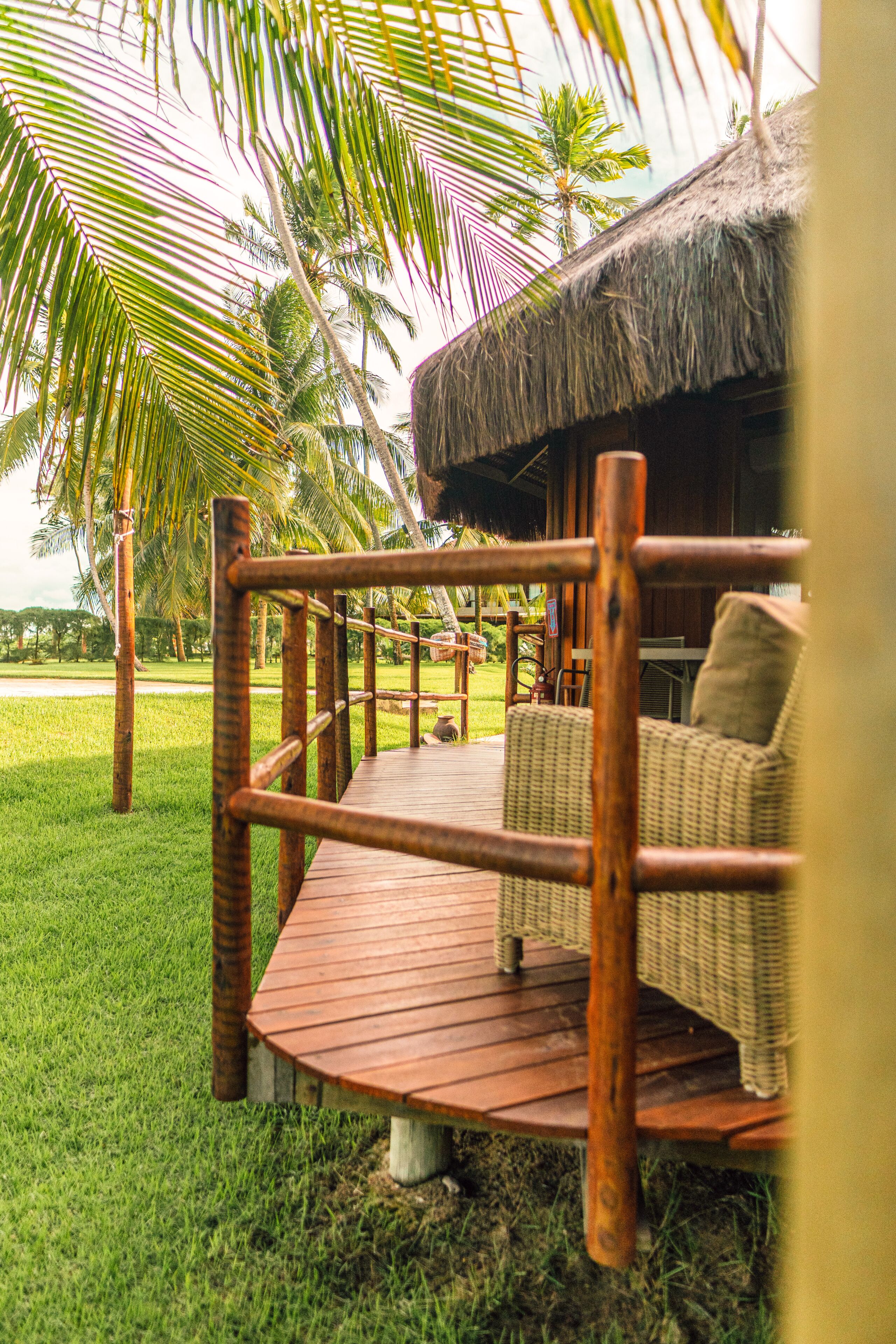 Una terraza de madera con una silla de mimbre, rodeada de palmeras y una estructura con techo de paja.