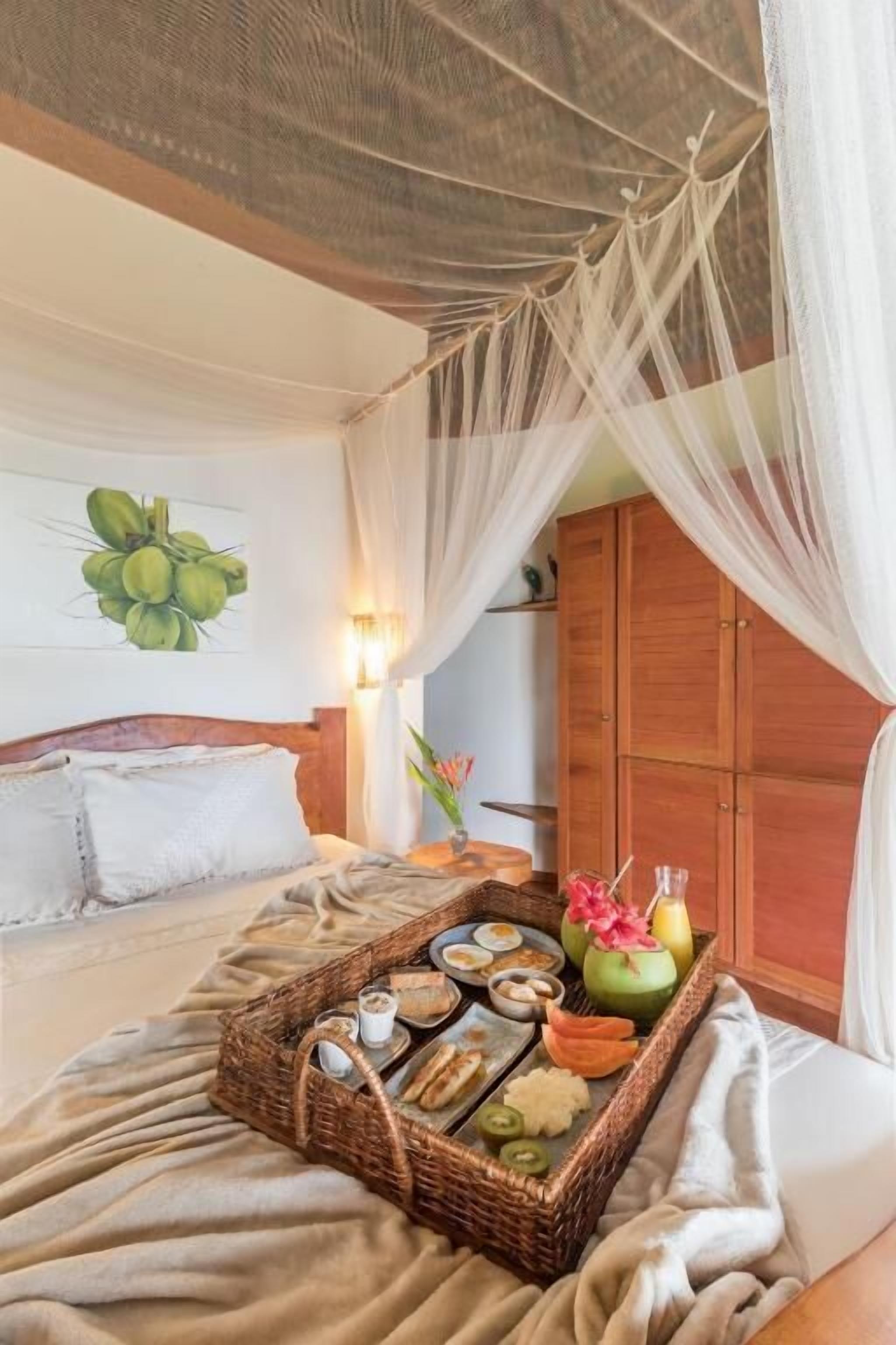 Una camera da letto con un letto, un comodino, un armadio in legno, un vassoio di vimini con cibo e bevande e un quadro raffigurante dei cocco.