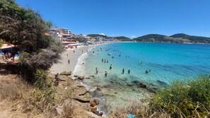 A beach with clear turquoise water, rocky shoreline, and buildings in the background. - Beachfront Apartment, Sea View, Garage, 2 Bedrooms, WI-FI, Cable TV, NETFLIX etc. (Arraial do Cabo)