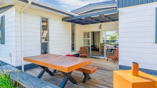 Una casa moderna con un patio que tiene una mesa y sillas de madera, una chimenea y un ventanal de vidrio que da a una cocina interior.