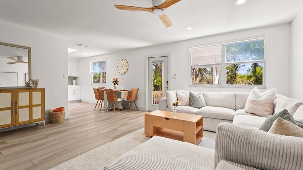 A spacious living room with a large window, a ceiling fan, and a dining area with a wooden table and chairs.