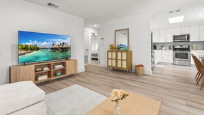 A modern living room with a flat-screen TV, wooden entertainment unit, and a dining area with a vase of flowers.
