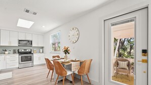 A modern kitchen with white cabinets, a microwave, and an oven. There is a dining area with a table, chairs, and a clock on the wall.