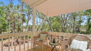 A covered outdoor seating area with wicker furniture and a small table, surrounded by trees.