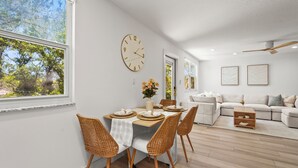 A modern living room with a dining area, featuring a large window, a ceiling fan, and a sofa set.