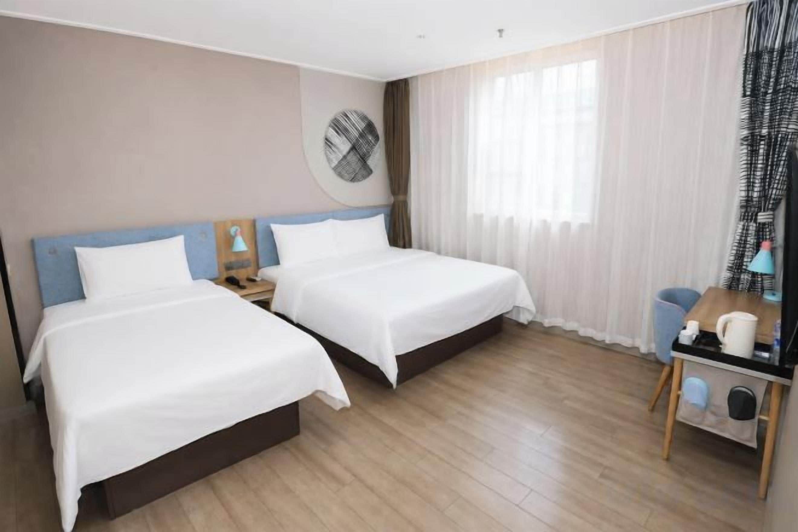 Una habitación de hotel con dos camas, un reloj de pared redondo, una mesita con lámpara y una ventana con cortinas.