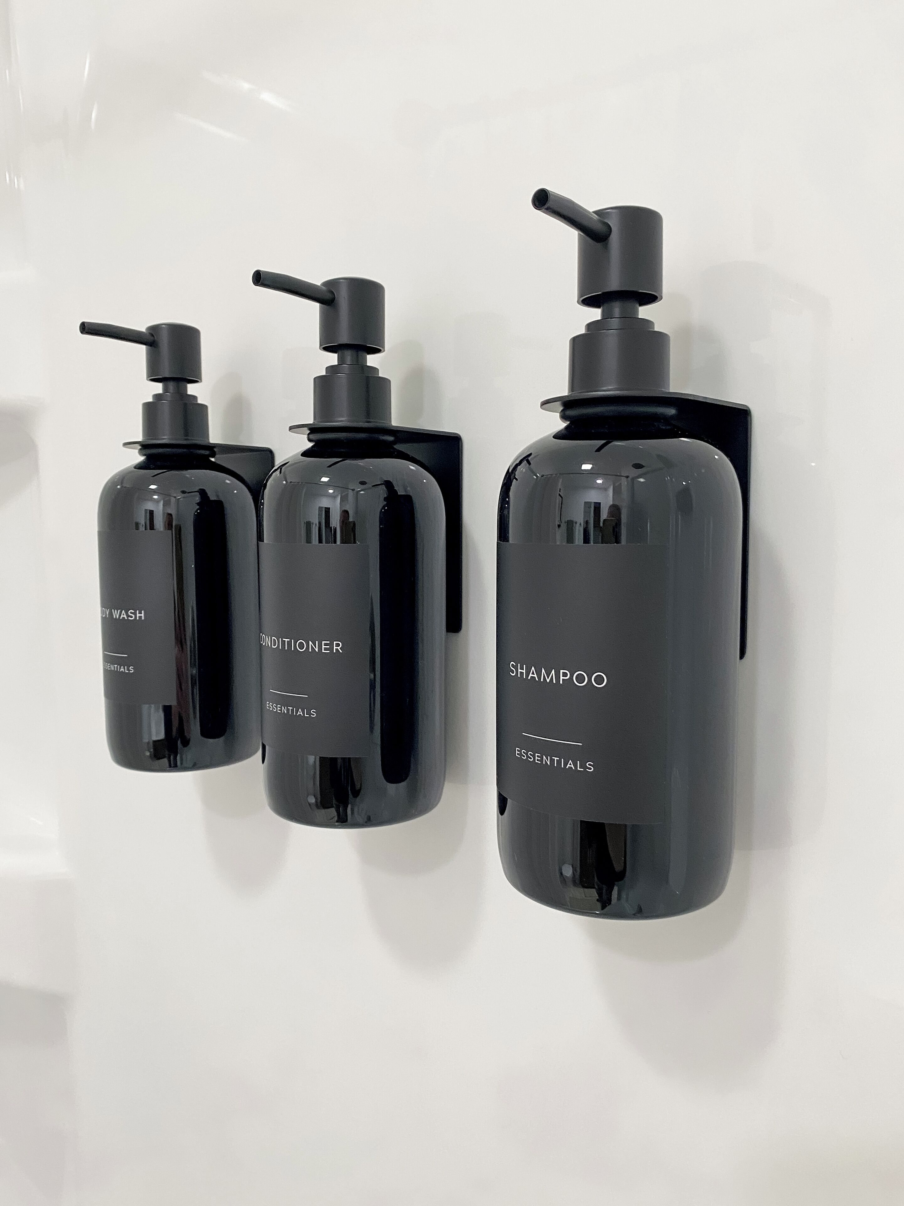 Trois flacons noirs de shampoing et d’après-shampoing sont fixés à un mur blanc.