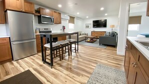 A modern kitchen with a stainless steel refrigerator, wooden cabinets, and a microwave. There is a dining area with stools and a sofa in the living room.