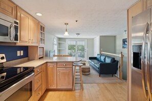 Een moderne keuken met houten kastjes, een centraal eiland, roestvrijstalen apparatuur en een zithoek met een zwarte bank en een poef.