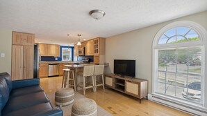 Een moderne woonkamer met een blauwe bank, rieten poufs, een televisie en een groot raam met zonwering.