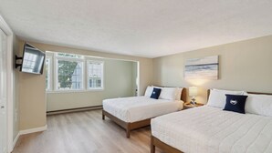 Een slaapkamer met twee bedden, een aan de muur bevestigde televisie, een raam met jaloezieën en een schilderij.