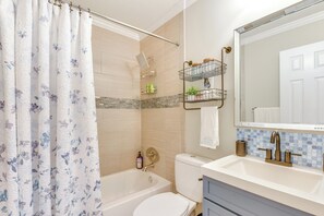 Un baño con bañera, inodoro y lavabo. Hay una cortina de ducha con estampado floral, un perchero y un espejo sobre el lavabo.