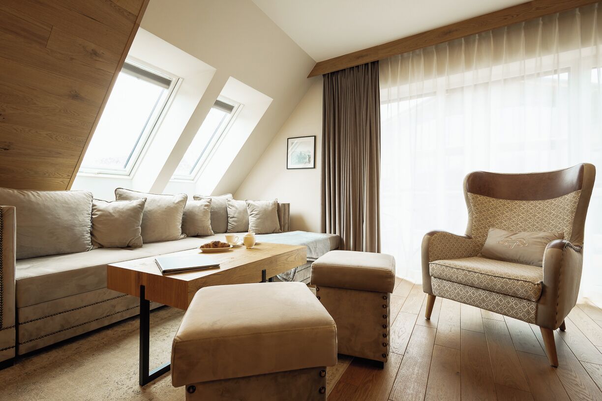 Sala de estar moderna com sofá, banqueta e poltrona, piso de madeira e grandes janelas com cortinas.