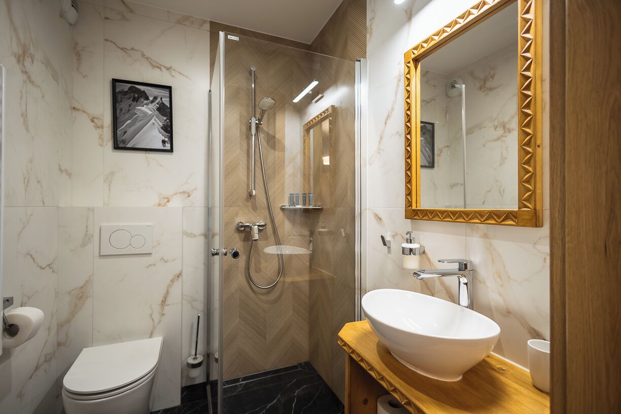 Une salle de bain moderne avec une douche à parois de verre, des toilettes blanches, un mur en marbre et un meuble-lavabo en bois surmonté d’un grand miroir.