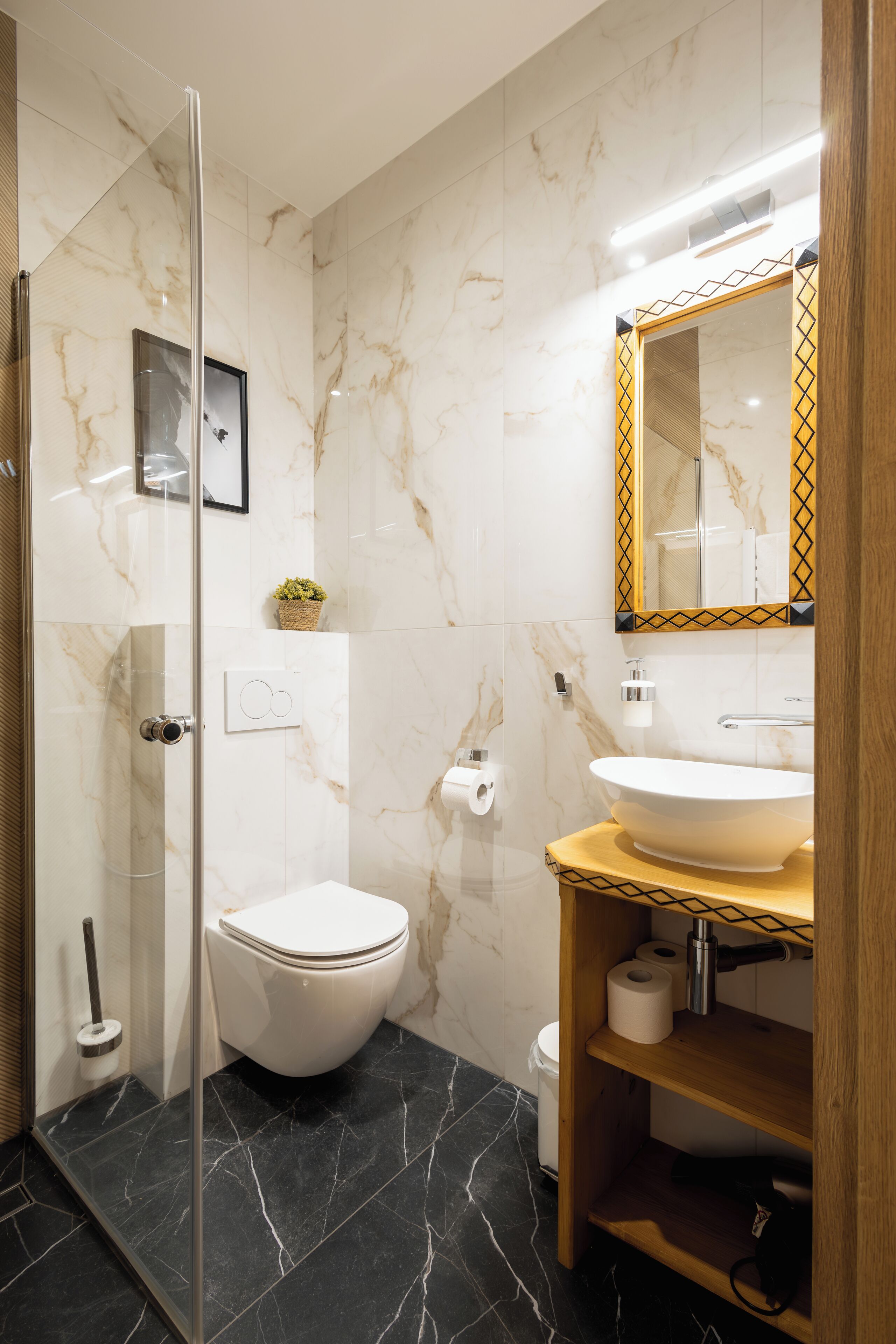 A modern bathroom with a glass shower enclosure, a white toilet, a marble-tiled wall, a wooden vanity with a sink, and a mirror with a decorative frame.