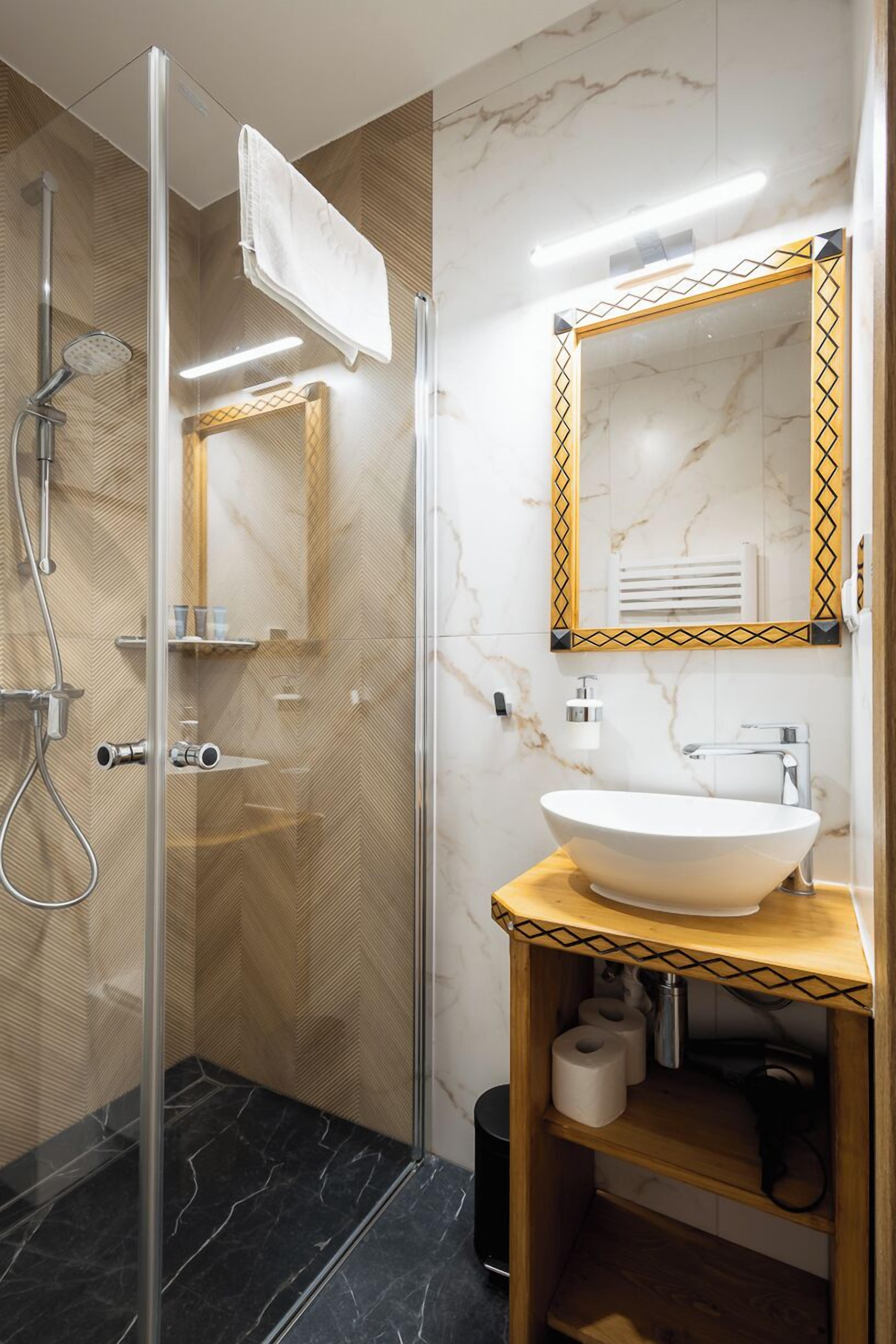 A modern bathroom with a glass shower enclosure, a marble-tiled wall, a white sink, and a wooden vanity.