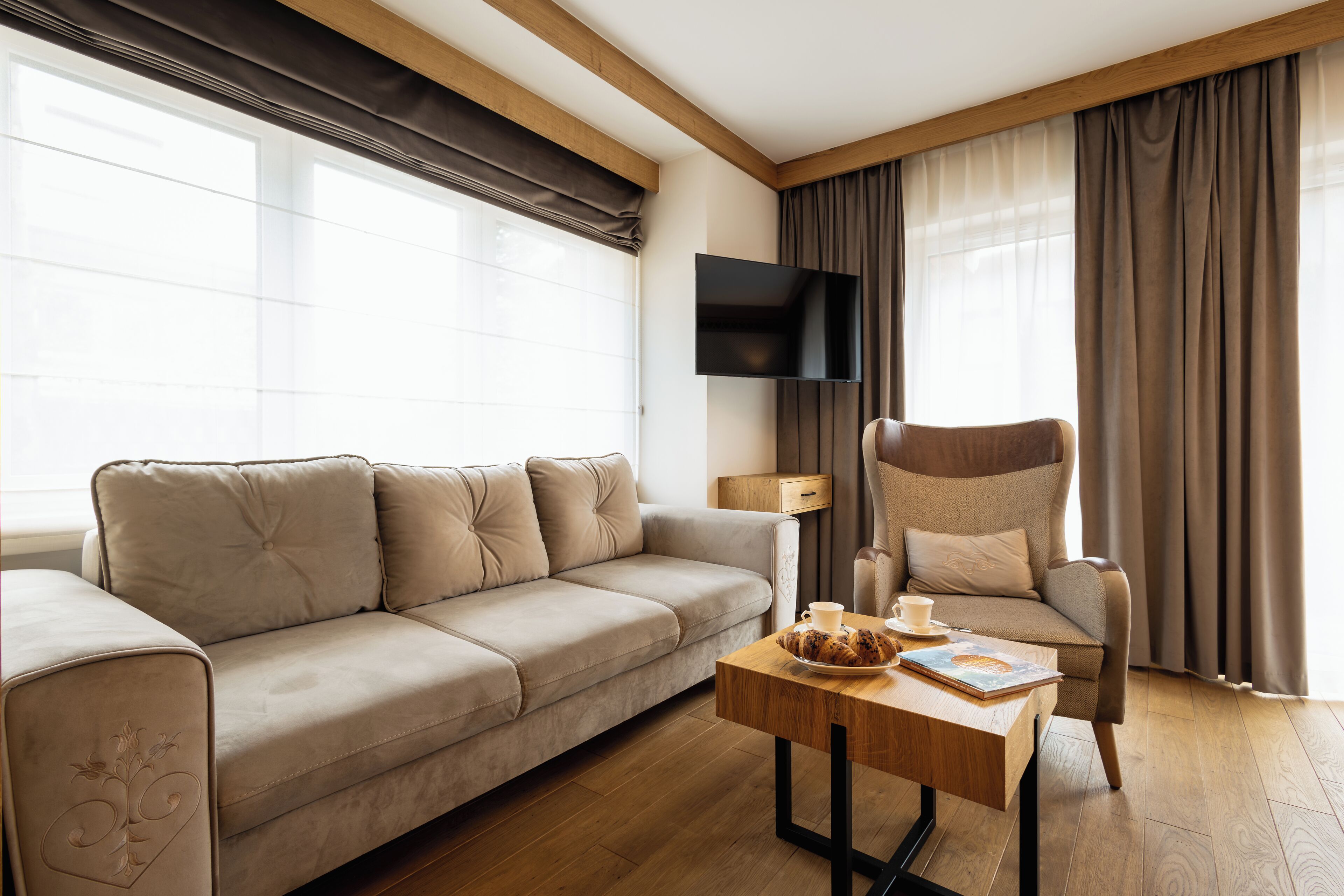 Un soggiorno moderno con un divano color beige, un tavolino in legno, una poltrona, una TV a schermo piatto e ampie finestre con tende.