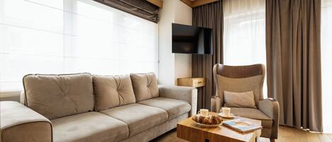 A modern living room with a beige sofa, a wooden coffee table, a chair, a flat-screen TV, and large windows with curtains.