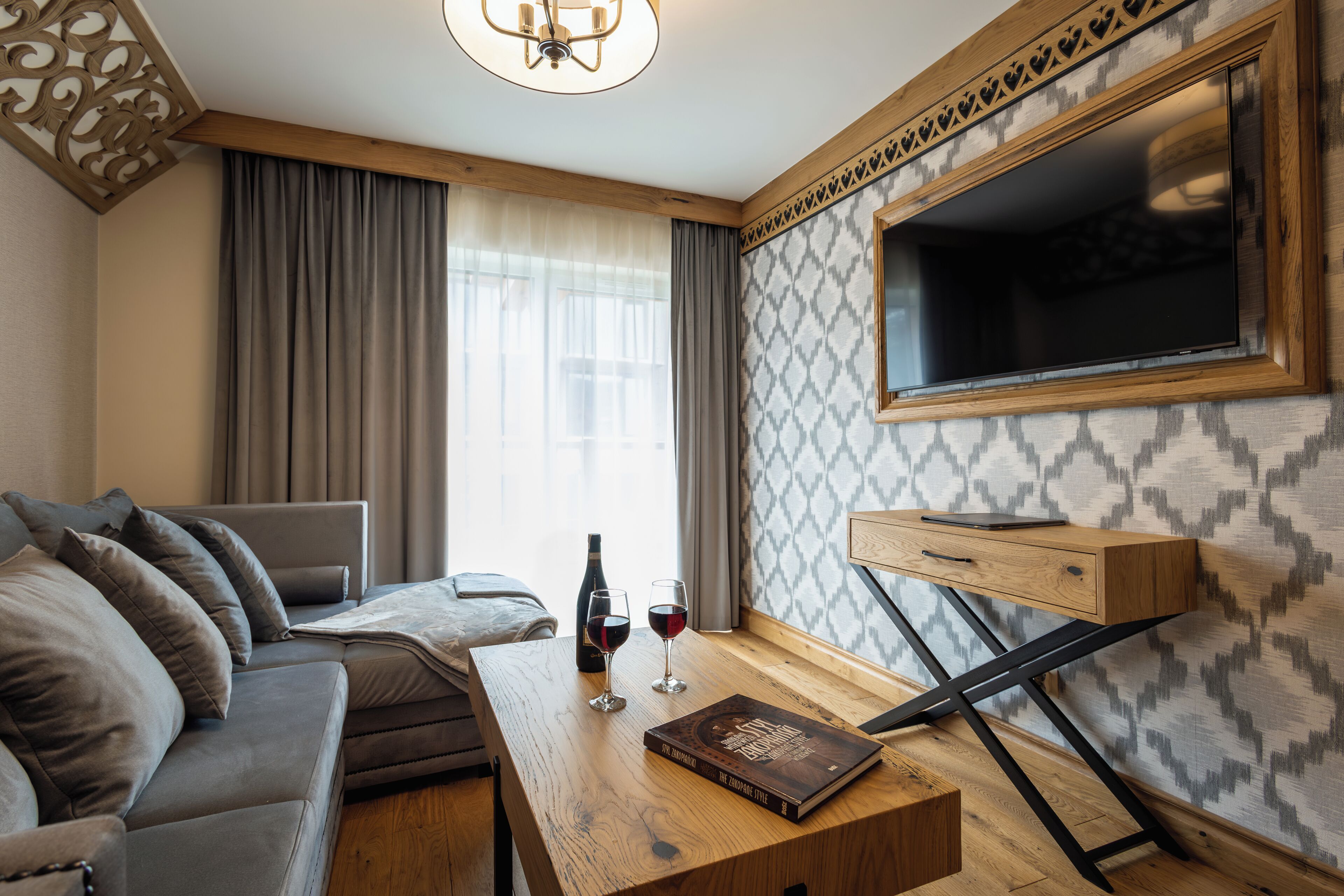 A modern living room with a sofa, a wooden coffee table, a TV mounted on the wall, and a patterned wallpaper.