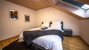 Ein Schlafzimmer mit schrägen Wänden, einem Bett mit weißer Bettwäsche, einem dunklen Kopfteil, einem Nachttisch und zwei gerahmten Bildern an der Wand.