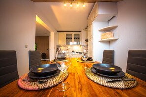 Un coin repas avec une table en bois dressée pour deux personnes, deux assiettes noires, deux verres à vin et une lampe en arrière-plan.