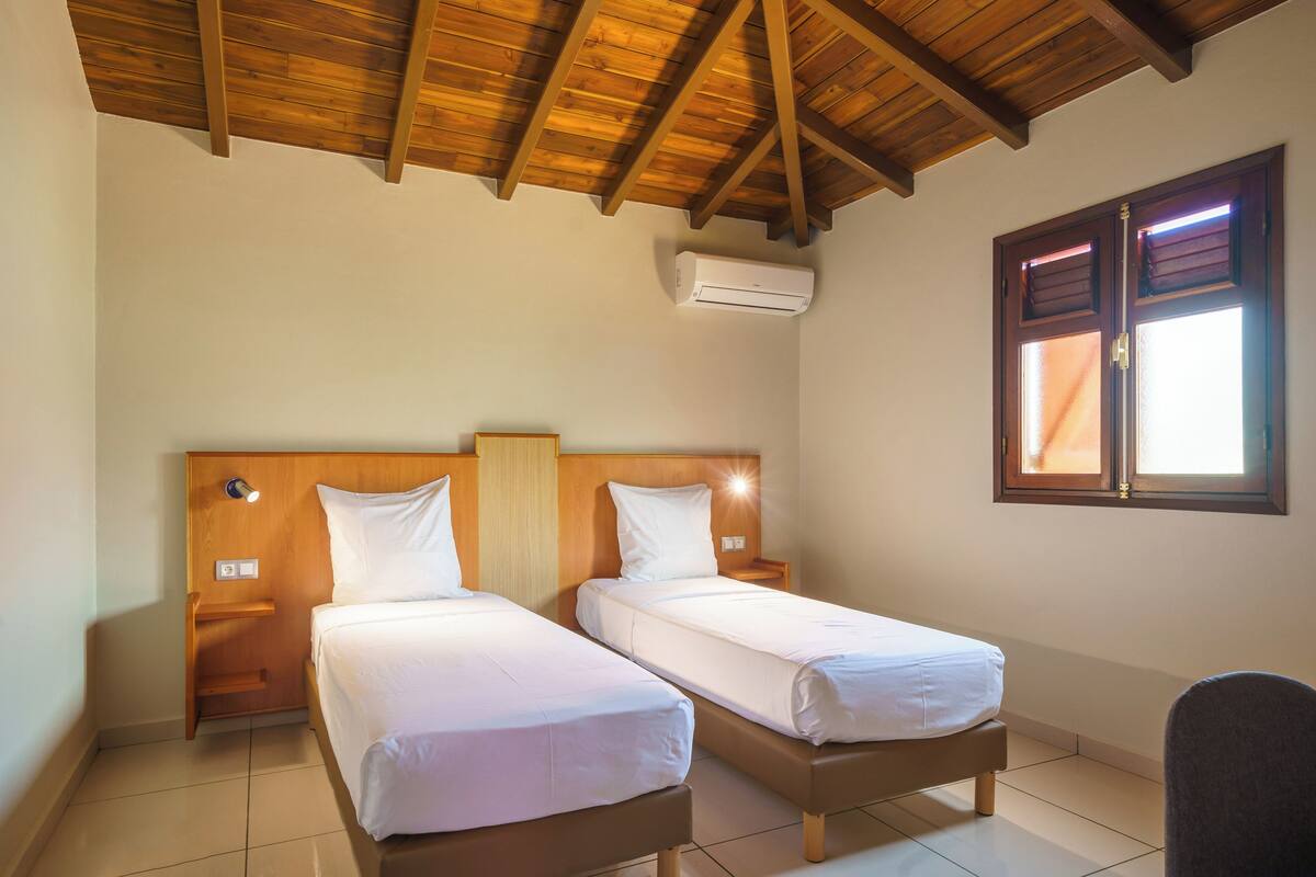 A room with two single beds, wooden headboards, a ceiling fan, an air conditioning unit, and a window with blinds.