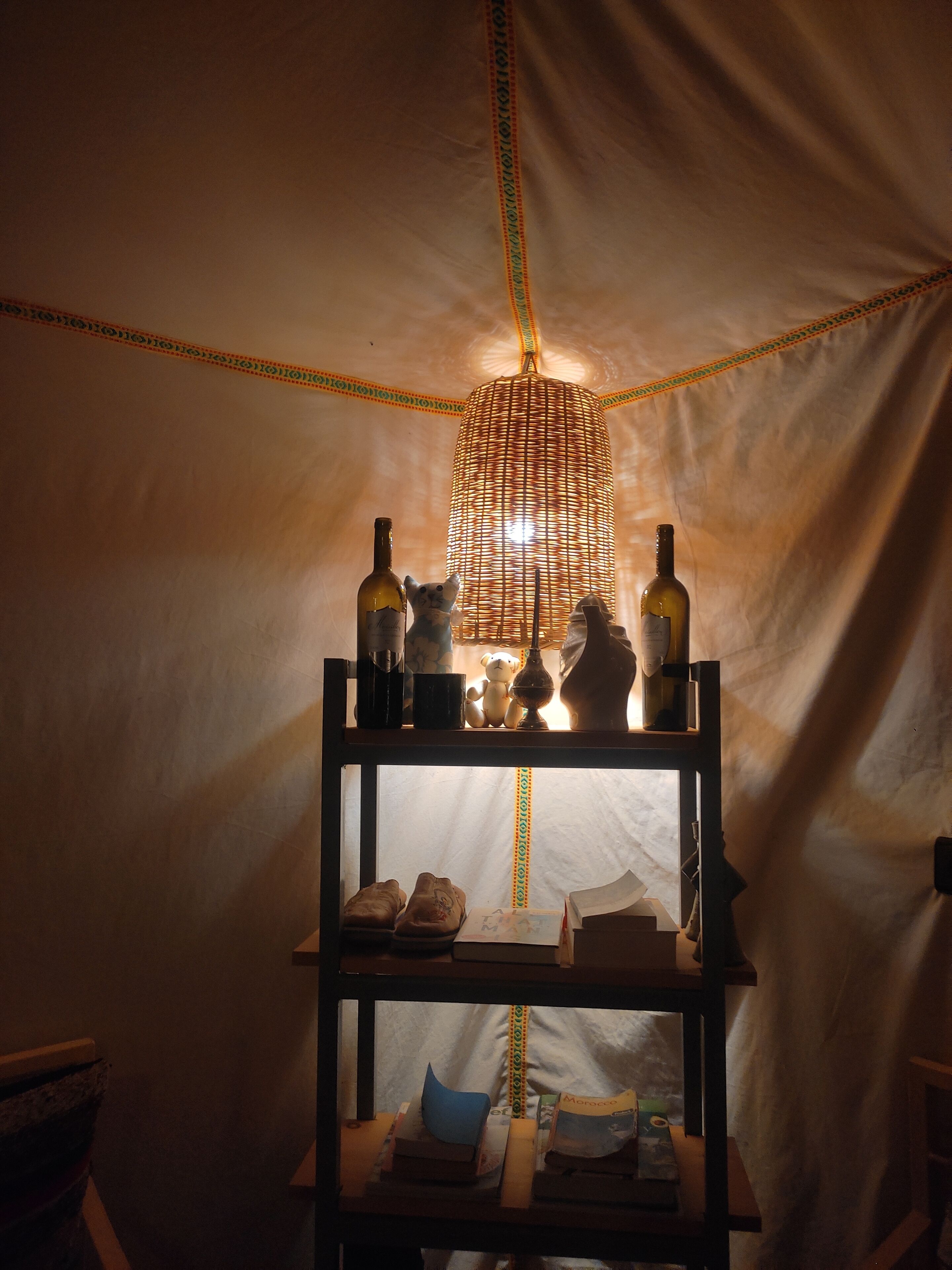 En hylde med flasker, en bamse og bøger i et teltlignende rum med en vævet lampe.