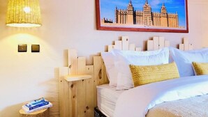 Een hotelkamer met een houten hoofdbord, een bed met wit en gouden beddengoed, een hanglamp, een lijst met een afbeelding van een gebouw, en een klein nachtkastje met een boek.