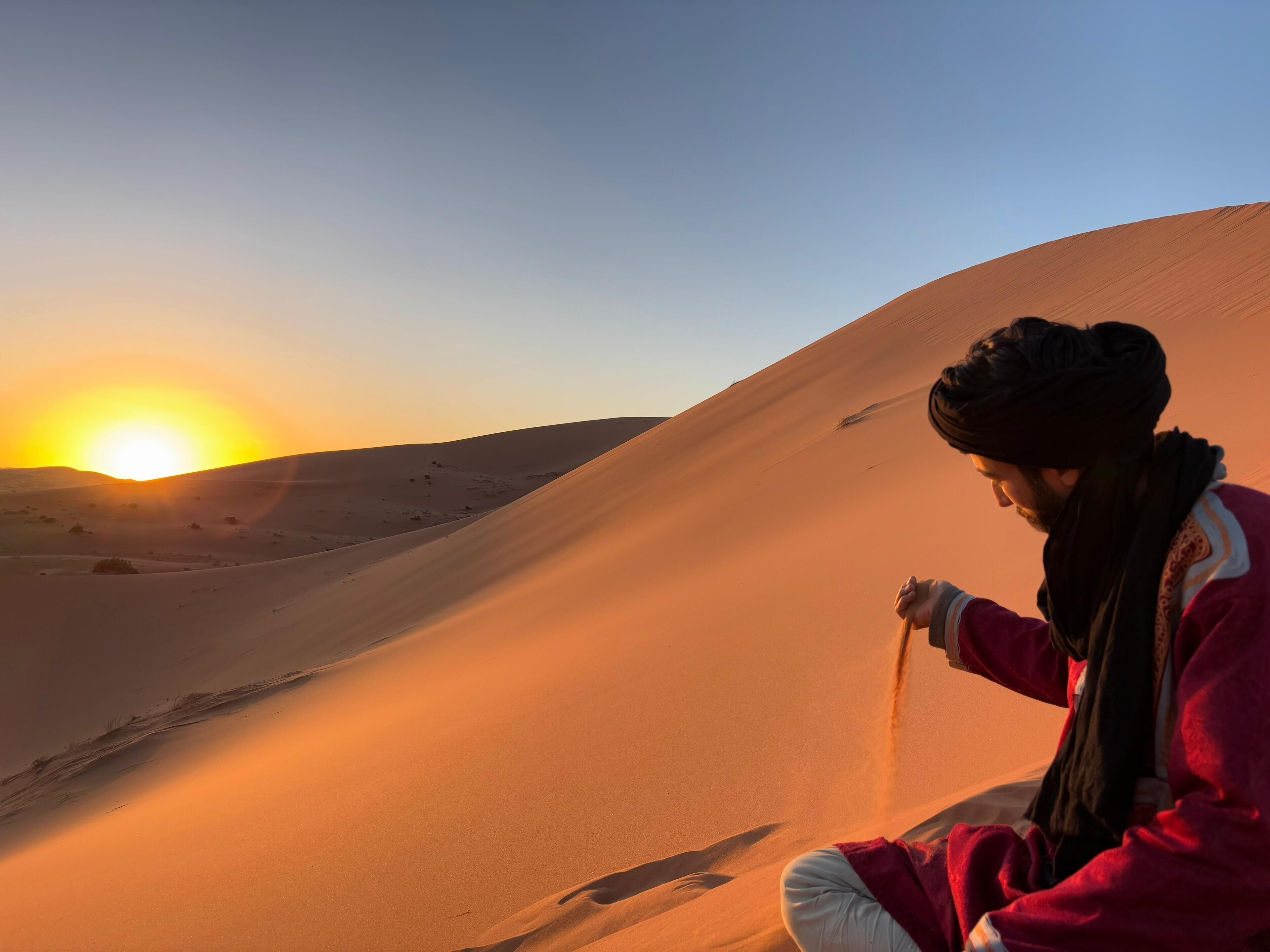 Una persona sentada sobre una duna de arena con un atardecer de fondo.