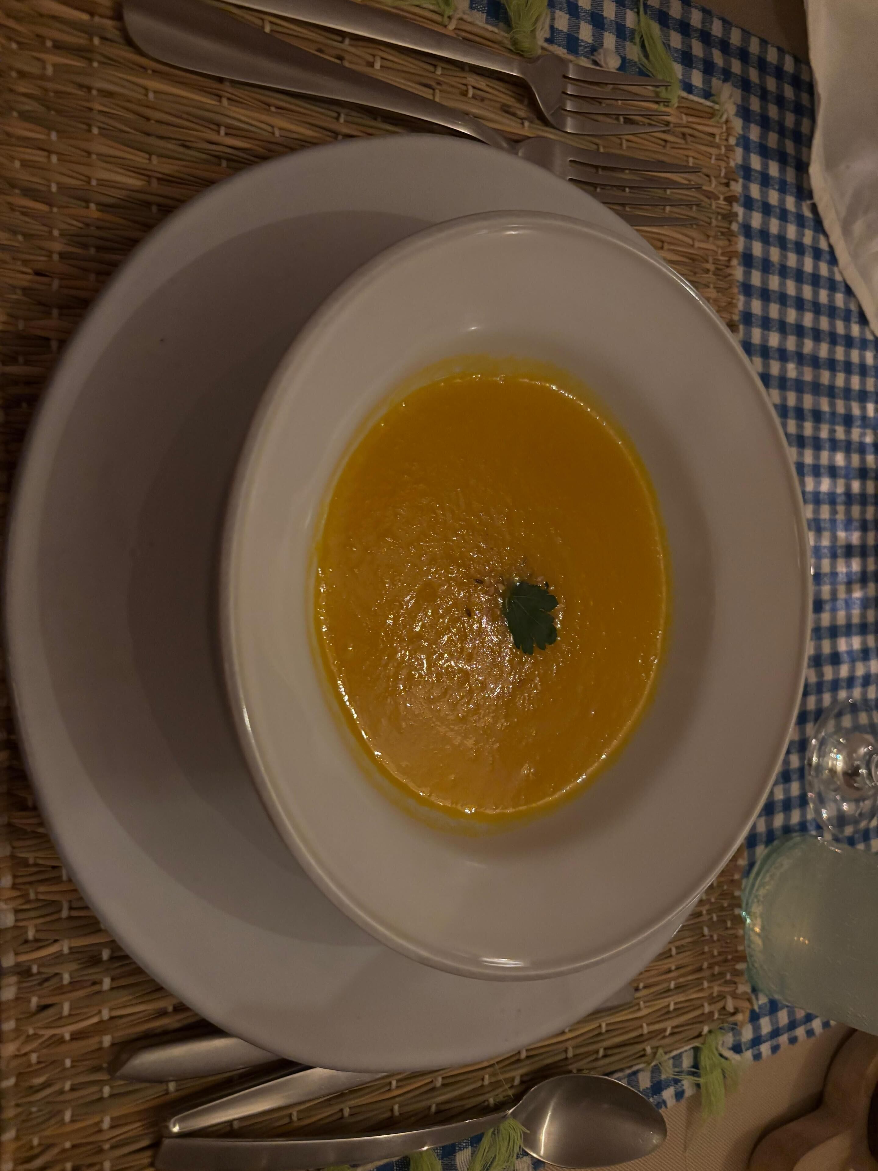 A bowl of soup on a white plate, placed on a checkered tablecloth.