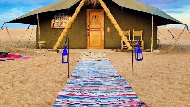 Een tent in de woestijn met een centrale deur, een kleurrijk geweven kleed dat ernaartoe leidt, en twee lantaarns aan weerszijden.