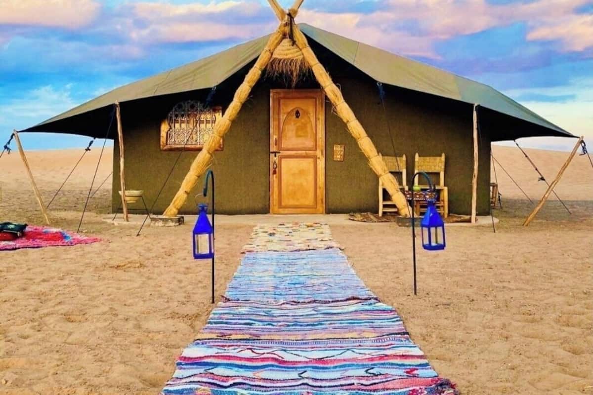 Une tente de désert dotée d’une porte centrale, d’un tapis coloré tissé qui mène à cette porte, et de deux lanternes placées de chaque côté.