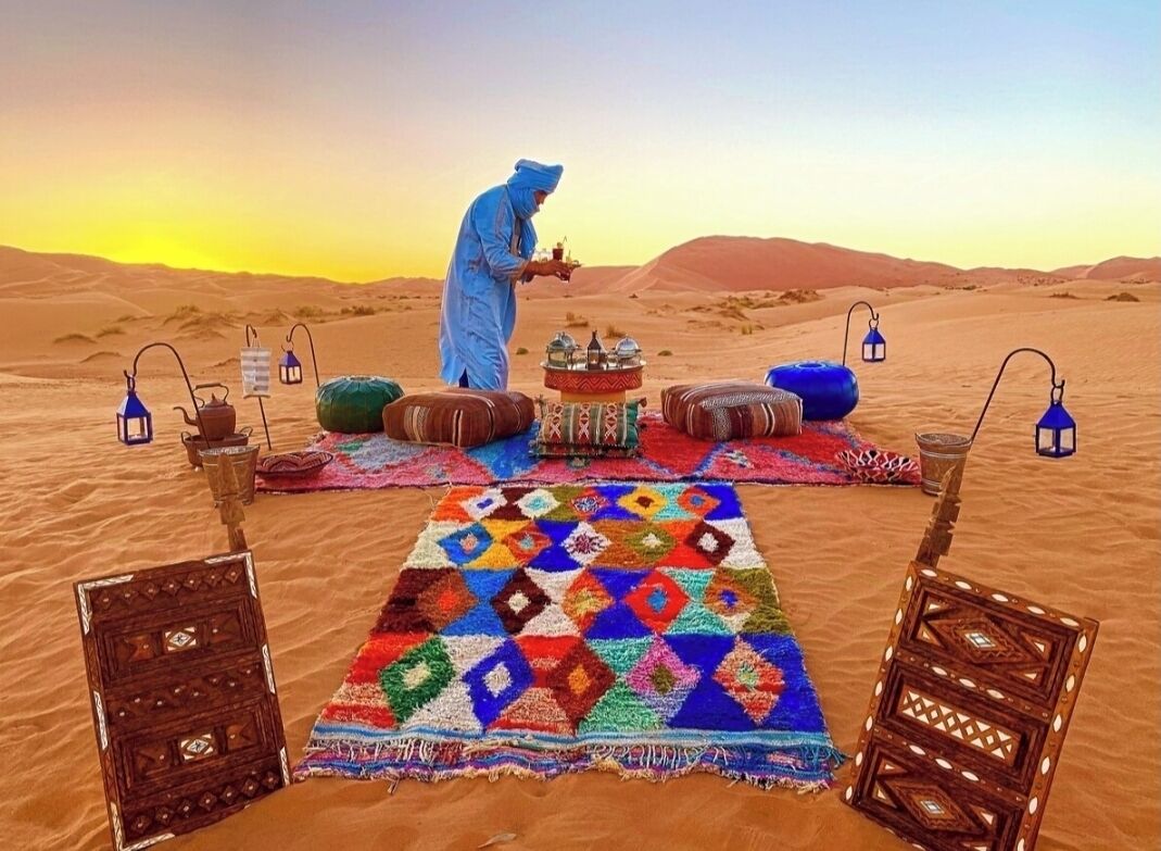 A desert scene with a person in traditional attire, a colorful rug, and a table with items.