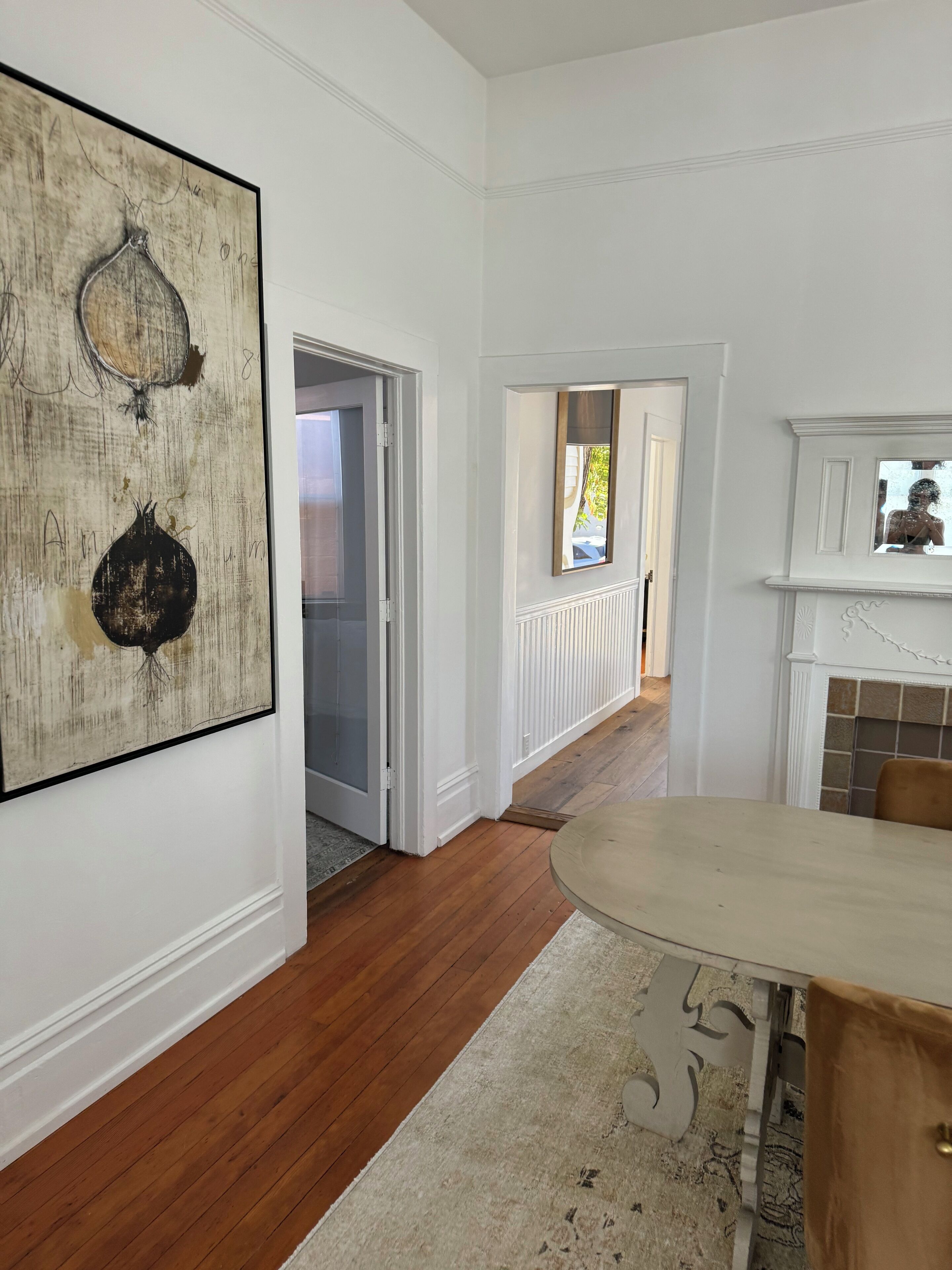 A hallway with wooden flooring, a white wall with a large abstract painting, and a door leading to another room.