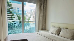 Une chambre d’hôtel avec un grand lit, une vue sur les immeubles et les palmiers depuis la fenêtre, et un balcon donnant sur la ville.
