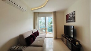 Un salon moderne avec un canapé, un téléviseur à écran plat et un balcon offrant une vue sur la verdure et les bâtiments.