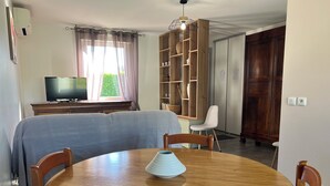 Un coin repas comprenant une table ronde en bois, quatre chaises en bois, un téléviseur posé sur un meuble et une bibliothèque avec des étagères en bois.